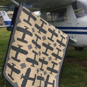 Puede incluir: Un edred&oacute;n beige y azul marino con un patr&oacute;n repetido de aviones. El edred&oacute;n est&aacute; sobre el ala de un avi&oacute;n peque&ntilde;o sobre hierba. El borde del edred&oacute;n es azul oscuro con un patr&oacute;n de puntos. Aproximadamente 150cm x 200cm.