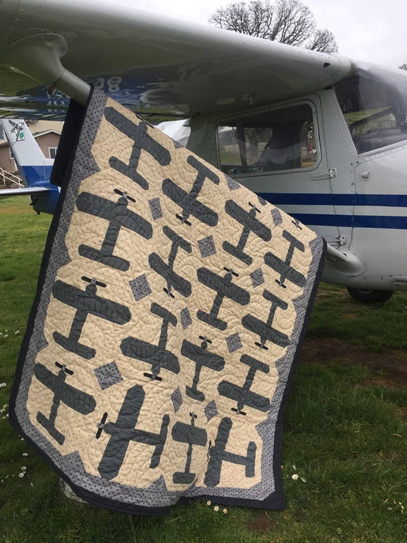 Puede incluir: Un edred&oacute;n beige y azul marino con un patr&oacute;n repetido de aviones. El edred&oacute;n est&aacute; sobre el ala de un avi&oacute;n peque&ntilde;o sobre hierba. El borde del edred&oacute;n es azul oscuro con un patr&oacute;n de puntos. Aproximadamente 150cm x 200cm.