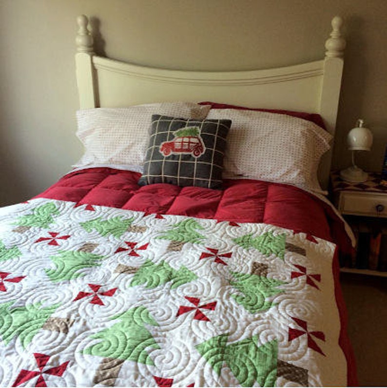 May include: A bed with a white headboard, red comforter, and a quilt featuring green trees and red accents. A decorative pillow with a car carrying a Christmas tree sits atop the bed, which also has white patterned pillows.