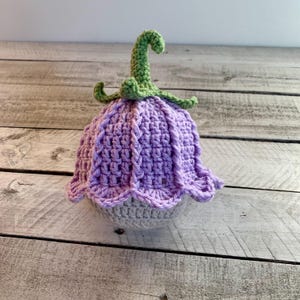 May include: A handmade crochet flower in shades of purple, green, and gray. The flower has a bell-shaped head with a scalloped edge, a green stem, and a gray base. The crochet work is detailed and textured.