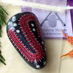 May include: Black painted stone with red and white decorative pattern. The stone has a teardrop shape and is decorated with a series of red lines and white star-shaped accents. The stone is displayed with a sprig of green foliage and a dried orange leaf.