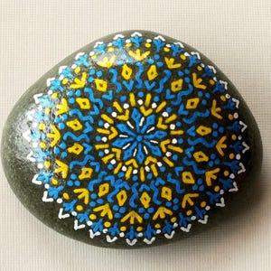 Könnte beinhalten: Ein handbemalter, dunkelgrauer Stein mit einem Mandala-Design. Das Mandala weist ein symmetrisches Muster in Blau, Gelb und Weiß auf. Das Design ist auf dem Stein zentriert und bedeckt den größten Teil seiner Oberfläche. Der Stein ist glatt und oval geformt.