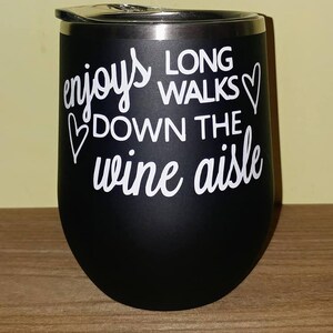 May include: Black stainless steel wine tumbler with a white graphic that reads "enjoys long walks down the wine aisle" with a heart symbol on either side of the text.
