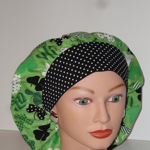 May include: A green and black patterned bouffant cap with a black polka dot band. The cap features a repeating pattern of shamrocks and other shapes. The cap is designed to cover the hair.