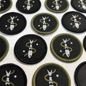 May include: A collection of black circular patches with a white fish design intertwined with a green snake-like symbol. Each patch has a green border and white dots. The patches are arranged on a white surface.