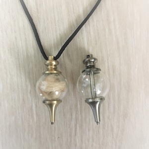 May include: Two gold-toned pendants on a black cord. The pendant on the left contains a small amount of light brown hair. The pendant on the right is empty.