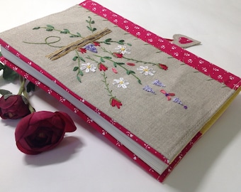 May include: A linen book cover with a floral embroidery design. The embroidery features pink, white, and purple flowers with green stems and leaves. The cover has a pink and white patterned border and a yellow edge.