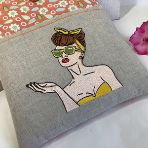 May include: A gray linen tote bag with a colorful embroidered image of a woman wearing sunglasses and a yellow headband. The woman is looking to the right and has her hand outstretched.