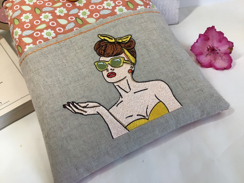 May include: A gray linen tote bag with a colorful embroidered image of a woman wearing sunglasses and a yellow headband. The woman is looking to the right and has her hand outstretched.
