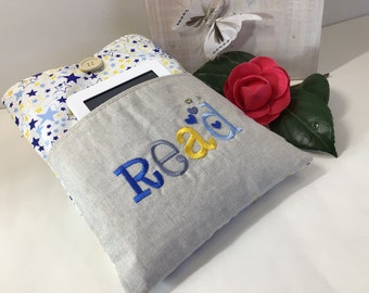 May include: A gray and white fabric book holder with a pocket for a tablet or book. The holder has a blue and yellow embroidered word "Read" and a blue and yellow star pattern on the back.