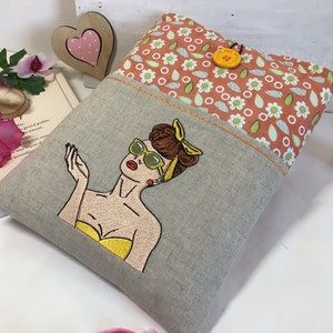 May include: A gray linen pillow with a pocket featuring a floral print in shades of orange and green. The pocket has a yellow button and an embroidered image of a woman with brown hair wearing sunglasses and a yellow top.
