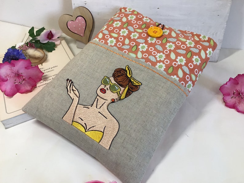 May include: A gray linen pillow with a pocket featuring a floral print in shades of orange and green. The pocket has a yellow button and an embroidered image of a woman with brown hair wearing sunglasses and a yellow top.