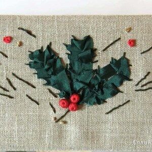 May include: A close-up of a green holly leaf embroidered on a beige linen fabric. The holly leaf is made of green ribbon and has red berries.