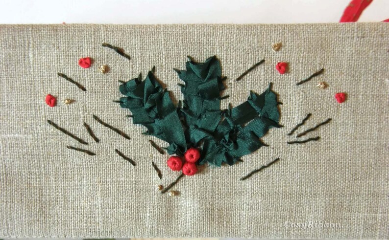 May include: A close-up of a green holly leaf embroidered on a beige linen fabric. The holly leaf is made of green ribbon and has red berries.