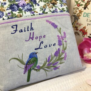 Może przedstawiać: Szara poduszka lniana z haftowanym wzorem lawendy i niebieskiego ptaka. Poduszka ma wyszyte słowa "Faith Hope Love".