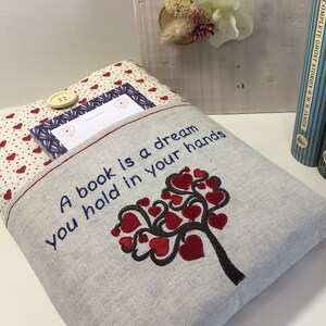 Puede incluir: Una funda de libro de lino gris con un dise&ntilde;o de &aacute;rbol de corazones rojos y el texto "Un libro es un sue&ntilde;o que tienes en tus manos". La funda tiene un bolsillo con estampado de corazones rojos y blancos en la parte delantera.