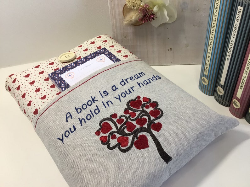 Puede incluir: Una funda de libro de lino gris con un dise&ntilde;o de &aacute;rbol de corazones rojos y el texto "Un libro es un sue&ntilde;o que tienes en tus manos". La funda tiene un bolsillo con estampado de corazones rojos y blancos en la parte delantera.