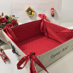 May include: A white fabric basket with red and gold star patterned lining and red ribbon handles. The basket is folded and has the text "Joyeux Noel" embroidered on the front.