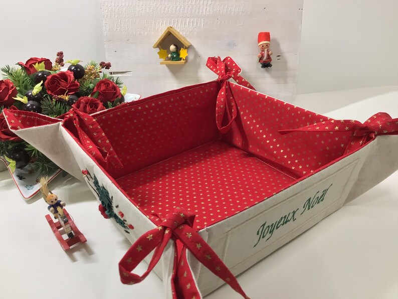 May include: A white fabric basket with red and gold star patterned lining and red ribbon handles. The basket is folded and has the text "Joyeux Noel" embroidered on the front.