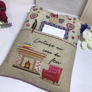 May include: Beige fabric book sleeve with an embroidered fireplace, chair, and text "Lecture au coin du feu." The top pocket has a floral pattern, a pen, and a small notepad. A wooden clock button secures the flap.