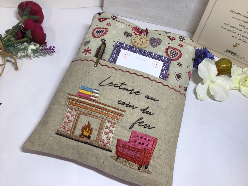 May include: Beige fabric book sleeve with an embroidered fireplace, chair, and text "Lecture au coin du feu." The top pocket has a floral pattern, a pen, and a small notepad. A wooden clock button secures the flap.