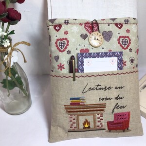 May include: A beige fabric book sleeve with a pocket, featuring a heart and floral pattern. It has a decorative clock button closure. The pocket holds a pen and a card. The sleeve is embroidered with a fireplace scene and the text "Lecture au coin du feu."