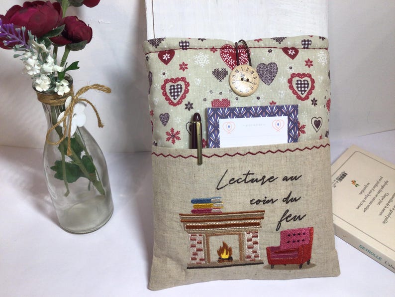 May include: A beige fabric book sleeve with a pocket, featuring a heart and floral pattern. It has a decorative clock button closure. The pocket holds a pen and a card. The sleeve is embroidered with a fireplace scene and the text "Lecture au coin du feu."