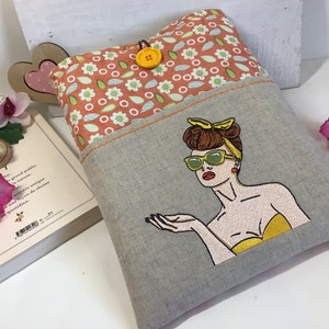 May include: A gray linen book cover with a pocket on the front. The pocket is lined with a peach fabric with a white floral pattern. The front of the cover features an embroidered image of a woman with brown hair, wearing sunglasses and a yellow top.