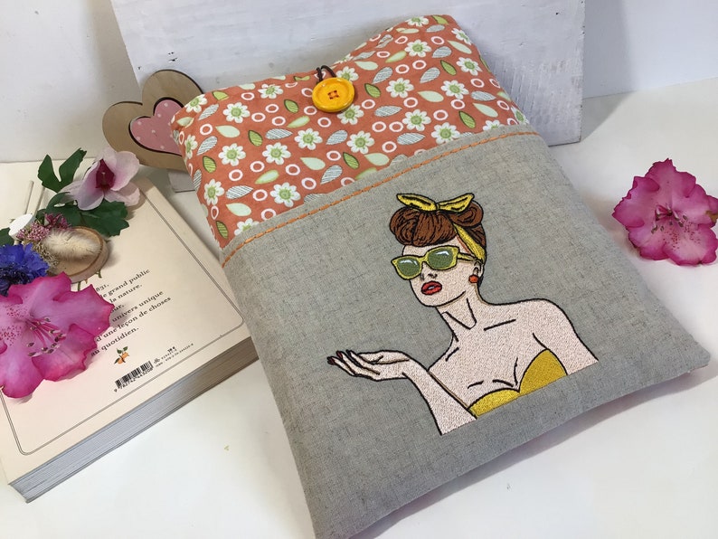 May include: A gray linen book cover with a pocket on the front. The pocket is lined with a peach fabric with a white floral pattern. The front of the cover features an embroidered image of a woman with brown hair, wearing sunglasses and a yellow top.