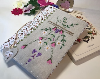 May include: A handmade book cover with a floral embroidery design. The cover is made of a light brown linen fabric with a white and brown floral pattern on the side. The embroidery features pink and purple flowers and green leaves. The cover is shown on top of a book with the word "Gallimard" printed on the spine.