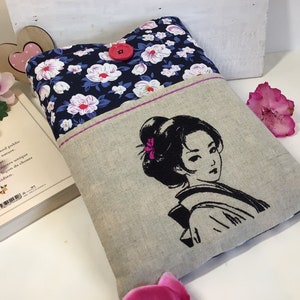 K&ouml;nnte beinhalten: Ein Leinen-Buchcover mit einem Blumenmuster auf der vorderen Klappe und einem gestickten Schwarzwei&szlig;bild einer Frau mit einer Blume im Haar auf der R&uuml;ckseite. Das Cover hat einen rosa Knopf.