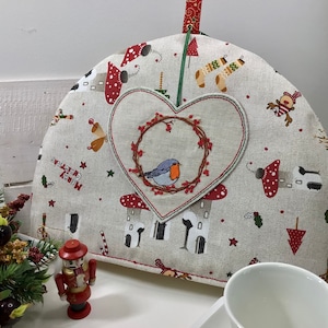 Puede incluir: Una calienta-tetera beige con un parche rojo en forma de corazón que presenta un pájaro azul en una corona. La calienta-tetera está decorada con un tema navideño, incluyendo setas, bastones de caramelo y estrellas.