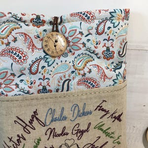 May include: A fabric book sleeve with a paisley pattern in shades of blue, orange, and red. The sleeve has a wooden button closure and is embroidered with author names. The sleeve is designed to protect books.