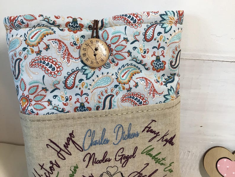 May include: A fabric book sleeve with a paisley pattern in shades of blue, orange, and red. The sleeve has a wooden button closure and is embroidered with author names. The sleeve is designed to protect books.