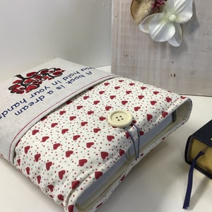 Puede incluir: Una funda para libro con fondo blanco y corazones rojos. La funda tiene una solapa con el texto "Un libro es un sue&ntilde;o que tienes en tus manos".