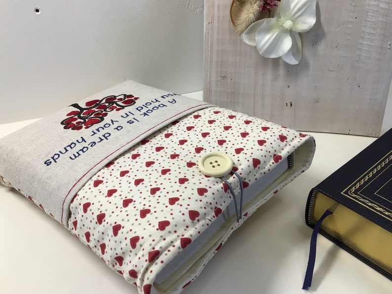 Puede incluir: Una funda para libro con fondo blanco y corazones rojos. La funda tiene una solapa con el texto "Un libro es un sue&ntilde;o que tienes en tus manos".