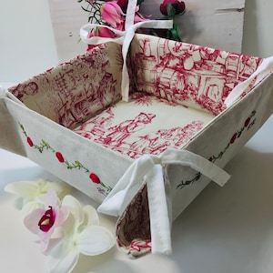 May include: A white linen bread basket with red floral embroidery and white ribbon ties. The basket is folded and has a red and white floral pattern on the inside.