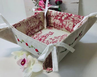 May include: A white linen bread basket with red floral embroidery and white ribbon ties. The basket is folded and has a red and white floral pattern on the inside.