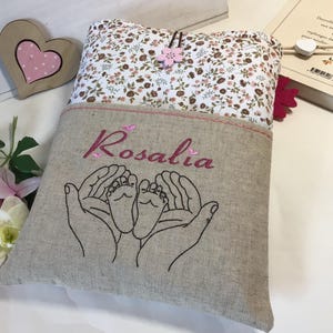May include: A personalized linen book cover with a floral pattern. The cover has a pocket with the name "Rosalia" embroidered in pink thread. The pocket also features an embroidered design of two hands holding a pair of baby feet.