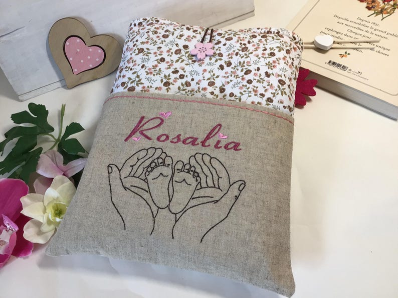 May include: A personalized linen book cover with a floral pattern. The cover has a pocket with the name "Rosalia" embroidered in pink thread. The pocket also features an embroidered design of two hands holding a pair of baby feet.