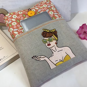 May include: A gray linen tablet sleeve with a pocket and a colorful embroidered design of a woman wearing sunglasses and a yellow top. The pocket has a floral patterned fabric lining.