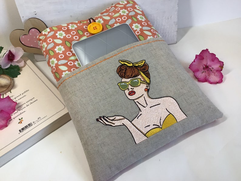 May include: A gray linen tablet sleeve with a pocket and a colorful embroidered design of a woman wearing sunglasses and a yellow top. The pocket has a floral patterned fabric lining.
