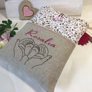May include: A beige linen book cover with a floral fabric pocket. The cover is embroidered with two hands holding a pair of baby feet and the name "Rosalia".