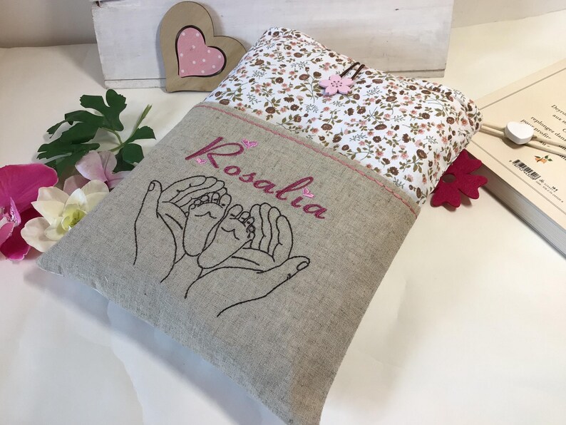 May include: A beige linen book cover with a floral fabric pocket. The cover is embroidered with two hands holding a pair of baby feet and the name "Rosalia".