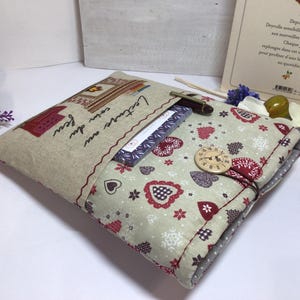 May include: A handmade book cover with a beige linen exterior and a patterned interior featuring red hearts and floral accents. The cover has a button closure and a pocket with the text "Lecture au coin du feu". A pen and a book are visible.