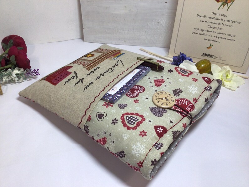 May include: A handmade book cover with a beige linen exterior and a patterned interior featuring red hearts and floral accents. The cover has a button closure and a pocket with the text "Lecture au coin du feu". A pen and a book are visible.