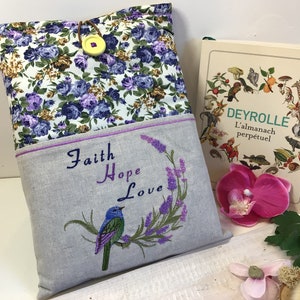 Może przedstawiać: Szara lniana okładka na książkę z kwiatowym wzorem i wyszytymi fioletowymi napisami "Faith Hope Love". Niebieski ptak siedzi na gałęzi lawendy. Okładka ma kieszeń z przodu z kwiatowym wzorem.