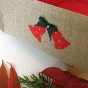 May include: A linen storage box with a red and gold star pattern and a green bow with a gold bell. The box is decorated with two red bells with gold stars.