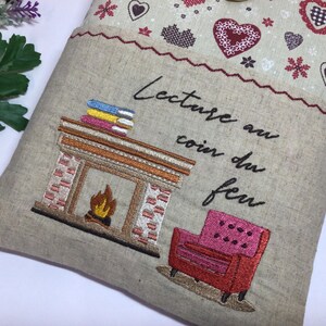May include: Embroidered pillow with a fireplace, books, and a pink armchair design. The pillow has the French text "Lecture au coin du feu" and is decorated with hearts and snowflakes.