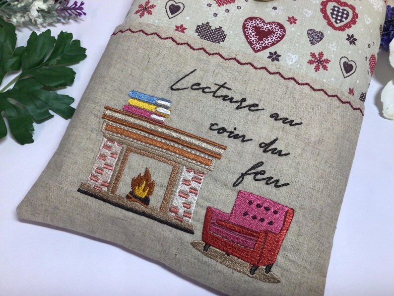 May include: Embroidered pillow with a fireplace, books, and a pink armchair design. The pillow has the French text "Lecture au coin du feu" and is decorated with hearts and snowflakes.